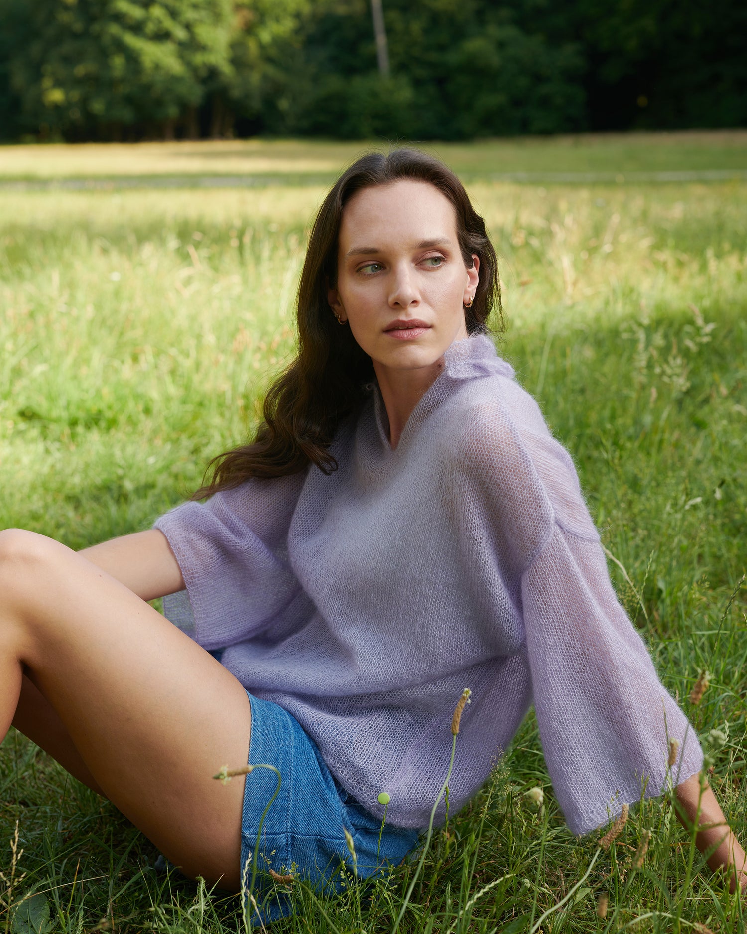 Mohair Seide Pullover FRIDA mit Rueschenkragen und Schlitz Seitenansicht offen an Model in Wiese sitzend in der Farbe lila nebel.
